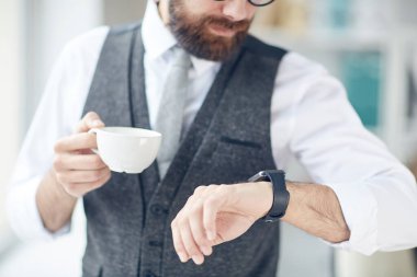 Gri yelekli şık iş adamının yakın çekimi fincandan kahve içmesi ve moladan önce kol saatine bakması.