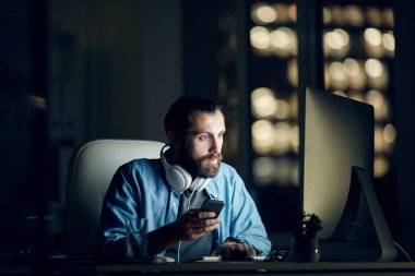 Kulaklıkları boynunda ciddi bir genç sakallı adam bilgisayarla masada oturuyor ve gece ofiste çalışırken telefon mesajlarını kontrol ediyor.