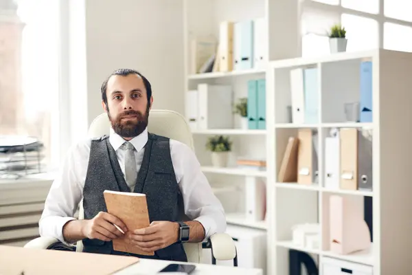Portrait of content successful young bearded manager in waistcoat and tie sitting at table in office and holding planner with pen