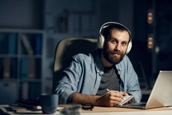 Portrait of content young bearded man in white headphones sitting at desk with laptop and making sketches in notepad while enjoying work at night