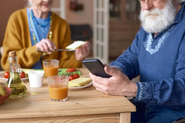 Close-up of senior man using smartphone while sitting at table and having lunch with his wife
