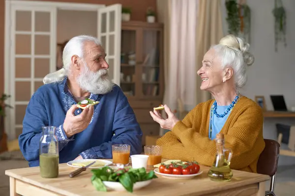 Senior couple eating sandwiches and talking to each other while having breakfast at home