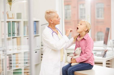 Klinikteki genç bir hastanın boğazını kontrol ederken çelik spatula kullanan laboratuvar önlüklü ciddi bir sarışın çocuk doktoru.