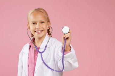 Pembe arka planda duran ve doktor olmak isterken steteskop kullanan laboratuvar önlüklü mutlu küçük kızın portresi.