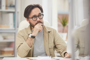 Ciddi düşünceli hippi Asyalı tasarımcı ofiste oturup web sitelerini incelerken monitörlere bakmaya odaklanmış.