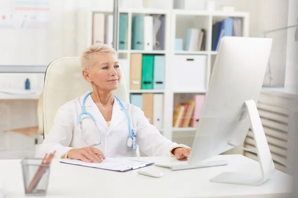 Content confident attractive lady doctor with short hair sitting at table and using computer while working with papers