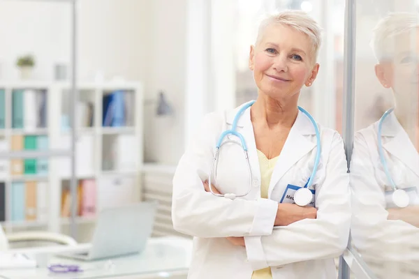 Portrait of content attractive lady doctor with short blond hair leaning on glassy wall in own office and crossing arms