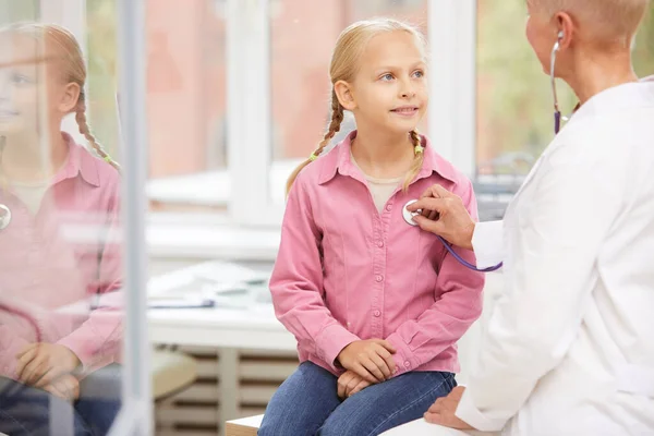 Schoolgirl visiting doctor: content pretty girl with braids sitting on examination table and looking at doctor while doctor listening to her lungs with stethoscope