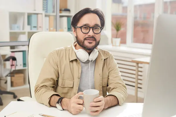 Portrait of content handsome young Asian manager with headphones on neck sitting at desk and drinking coffee in office