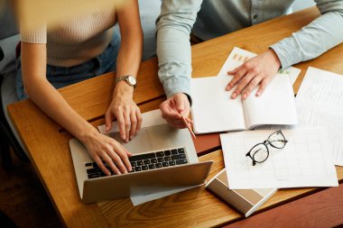 Ofisteki ahşap masada oturan ve verileri karşılaştırırken laptop ve kağıtları kullanan tanınmayan meslektaşların yüksek açılı görüntüsü