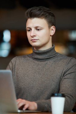 Boğazlı kazaklı genç web tasarımcısı kahve fincanı ile masada oturuyor ve bilgisayarla ofis dışında çalışıyor.