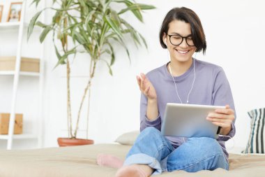 Gözlüklü, mutlu ve çekici bir kadın kanepede oturup el kol hareketi yaparken arkadaşınla internet üzerinden iletişim kurmak için tablet kullanıyor.
