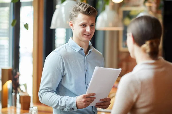 Content young male restaurant manager with papers standing in cafe and talking to waitress about customer service