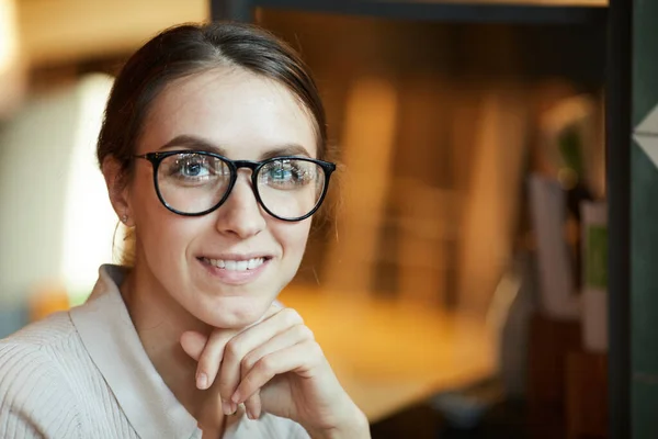 Positive young pretty lady with kind smile wearing stylish glasses leaning head on hand and looking at camera