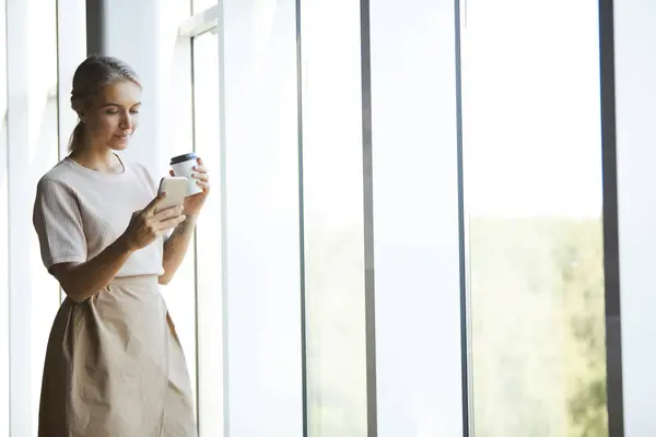 Content attractive blond woman in skirt standing by window and reading message on phone while drinking coffee