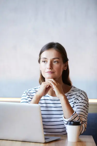 Serious pensive female internet marketer in casual clothing sitting at table in cafe and leaning head on hands while thinking of web design