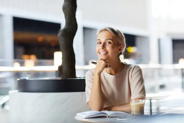 Portrait of cheerful beautiful young female freelancer in sweater sitting at table with planner and planning day in modern cafe