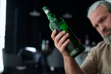 Elinde alkol şişesi olan sarhoş adam evde otururken içki içiyor.