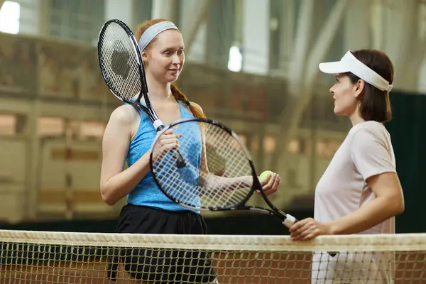 Content young female tennis players with racquets standing at net and discussing match together at modern indoor court