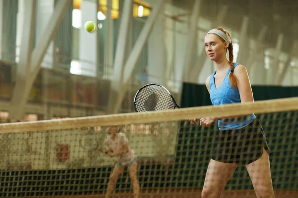 Content redhead woman in activewear standing by net on indoor court and holding racket with hands while playing tennis in team