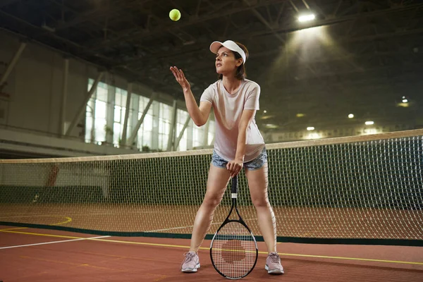 Content confident brunette woman in sportswear standing on indoor court and throwing tennis ball before serving