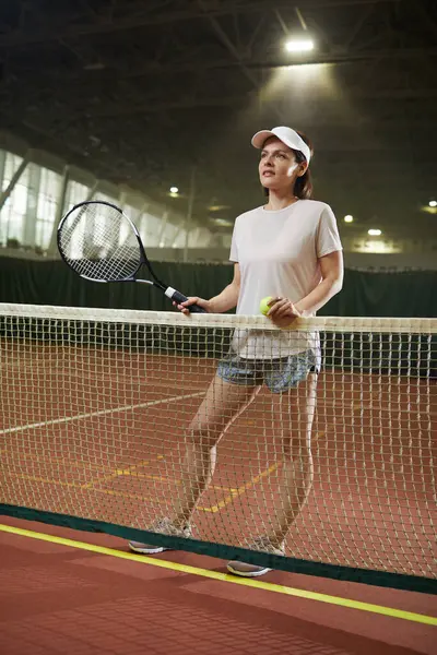 Smiling attractive female player in activewear standing by tennis net on court and holding racket and ball while talking to competitor