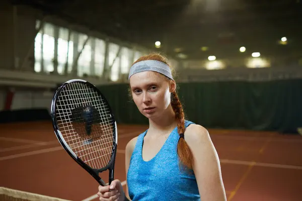 Portrait of serious sweaty young redhead woman in sports headband standing on contemporary tennis court and holding racket