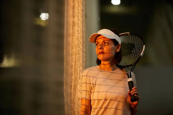 Serious brunette woman in cap standing by net and holding racket, she getting tense before big tennis match