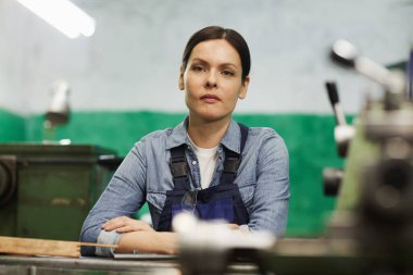 Fabrika atölyesinde masaya dayanmış, kahverengi saçlı, azimli kadın mühendisin portresi.