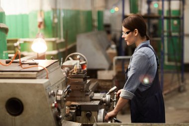 Odaklanmış orta yaşlı kadın endüstriyel işçi tulumları içinde elle torna tarama yaparken metal işlemleri yapıyor.