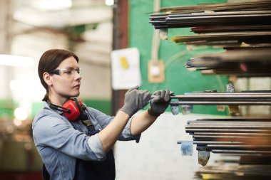 İşletme eldivenli, yoğun, genç esmer sanayi işçisi işlemek için metal çubuğu seçiyor.