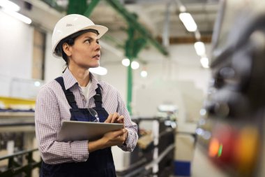 Fabrikada duran ve Cnc makinesini çalıştırmak için tablet kullanan çok düşünceli bir kadın teknik mühendis.