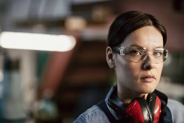 Portrait of serious attractive tough woman with brown hair wearing red ear protectors on neck standing at factory workshop