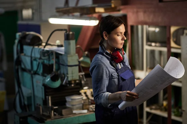 Serious emancipated brunette woman with ear protectors on neck standing by lathe and working with engineering sketch