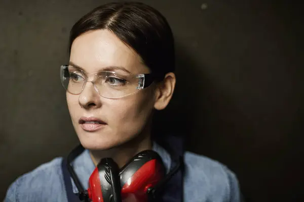 Close-up of smiling gritty industrial woman in safety goggles wearing ear protectors on neck standing against dark wall and looking aside
