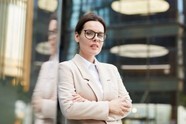 İş merkezinin lobisinde dikilirken göğsünde kollarını kavuşturan şık ceketli ve gözlüklü kendinden emin orta yaşlı kadının portresi.