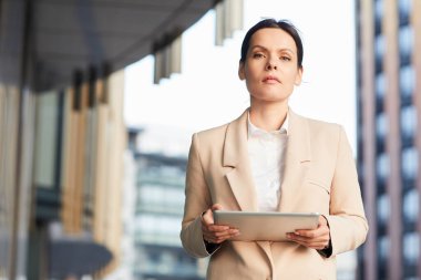 Kendine güveni tam orta yaşlı, şık ceketli bayan girişimcinin portresi iş bölgesinde duruyor ve tablet kullanıyor.