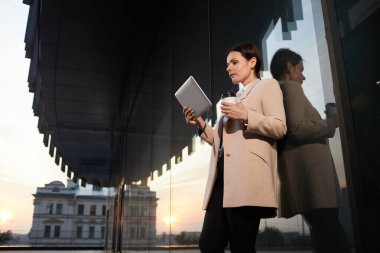 Duvara yaslanıp tablette video sunumu izleyen zarif ceketli, düşünceli, orta yaşlı, çekici bir kadın.