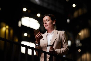 Düşünceli, çekici, orta yaşlı iş kadını kahve fincanı tırabzanda duruyor ve telefon notlarını analiz ediyor.