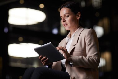 Gece vakti sosyal medyadan istatistik alırken tablet kullanan şık giyimli düşünceli orta yaşlı iş kadını.