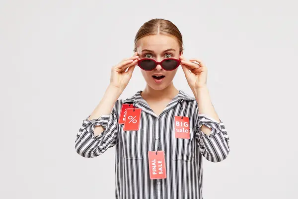 Surprised young woman in blouse with red Sale tags keeping mouth open and looking over sunglasses during Black Friday sale