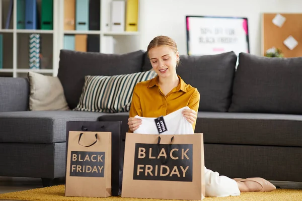 Content pensive young woman in blouse sitting on carpet at home and unpacking shopping bags with Black Friday lettering