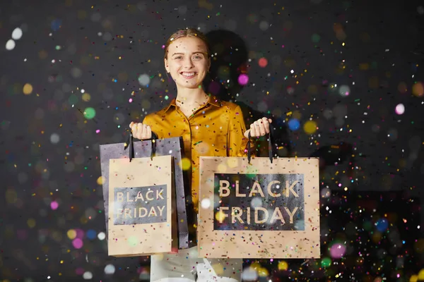 Happy excited lady shopper in silk shirt standing under confetti against black wall and holding paper bags with Black Friday text