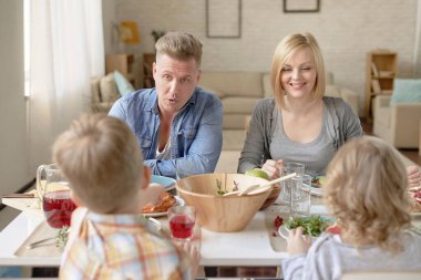 Çekici sarı saçlı beyaz çift yemek masasında çocuklarla oturuyor, iştah açıcı ev yemekleri yiyor, sohbet ediyor ve günün tadını çıkarıyorlar.