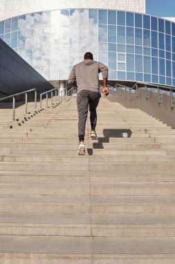 Tanınmayan atletik siyah adam çimento merdivenlerden çıkıp modern iş merkezine doğru koşuyor.