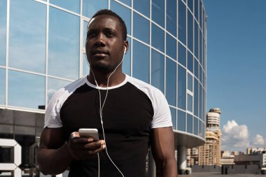 Akıllı telefonlu, modern ticari binaya karşı duran, kulaklıkla müzik dinleyen yakışıklı Afrikalı adamın düşük açılı görüntüsü.