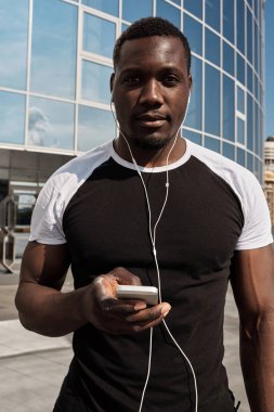 Akıllı telefonu ve kulaklığı olan, modern cam bina karşısında kameraya bakan yakışıklı Afrikalı Amerikalı bir adamın portresi.