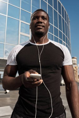Yakışıklı, atletik, akıllı telefonlu ve kulaklıkla müzik dinleyen siyahi bir adamın düşük açılı görüntüsü.