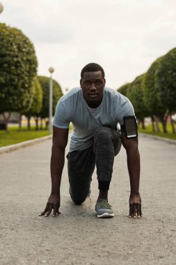 Sporcu koşmaya hazır. Atletik Afrikalı Amerikalı genç bir adamın portresi parktaki başlangıç pozisyonunda ve kameraya bakıyor.