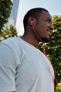 Genç kaslı yakışıklının düşük açılı görüntüsü. Dışarıda dikilen siyah adam, kırmızı kulaklıklarla müzik dinliyor ve mutlu bir şekilde gülümsüyor.